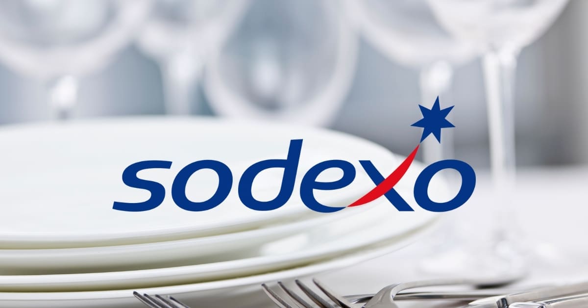 Logo de Sodexo apposé sur une pile d’assiettes blanches avec des verres flous en arrière-plan